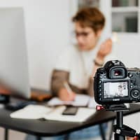 Unrecognizable blurred lady working at table in modern office against photo camera placed on tripod